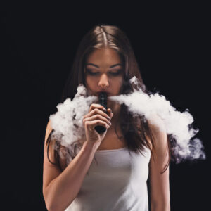 Young woman vaping, studio shot. Brunette girl blowing a cloud of smoke on black background. Nicotine free smoking and vapor concept, copy space